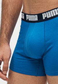 Крупен план на мъж, носещ сини боксерки Puma с черна талия, на която е лого на Puma в бяло.