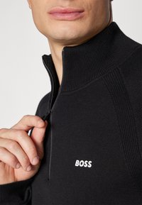 Črni pleten pulover z zadrgo in teksturiranimi rebrastimi detajli ter belim logotipom "BOSS" na prsih, prikaz natančnega pregleda detajlov.