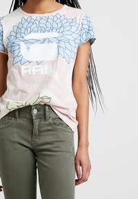 Žena v světlém růžovém tričku s velkým modrým květinovým grafikou a bílým textem "RAW", doplněná olivově zelenými kalhotami.