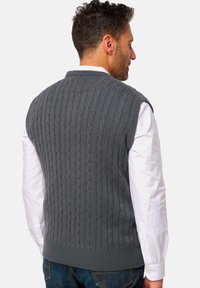 Graue Strickweste mit Zopfmuster, rundem Ausschnitt und geripptem Saum, getragen über einem weißen Langarmshirt und Jeans.