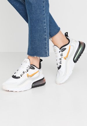 Persoon die witte Nike Air Max 270 React sneakers draagt met een oranje swoosh en zwarte details, gecombineerd met versleten blauwe spijkerbroek.