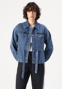 Veste en denim bleu moyen, à coupe décontractée, avec fermeture par boutons, poches poitrine et liens sur les côtés. Tissu texturé avec un col classique.