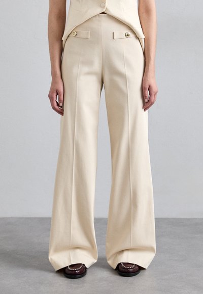 Pantalones de pierna ancha en color beige, confeccionados con una tela suave, con pliegues frontales y botones decorativos en los bolsillos. Corte a medida con líneas limpias.