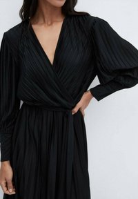 Une femme portant une robe portefeuille plissée noire avec de longues manches bouffantes et un décolleté en V profond, la main posée sur sa taille.