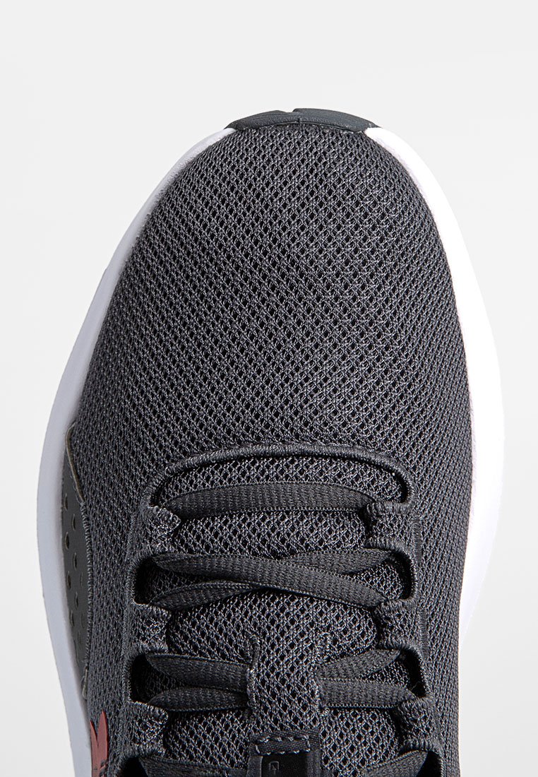 Zapatilla deportiva de color gris oscuro con una parte superior de malla texturizada, acentos en rojo contrastante y una suela blanca acolchada. Presenta cordones planos y lados perforados.