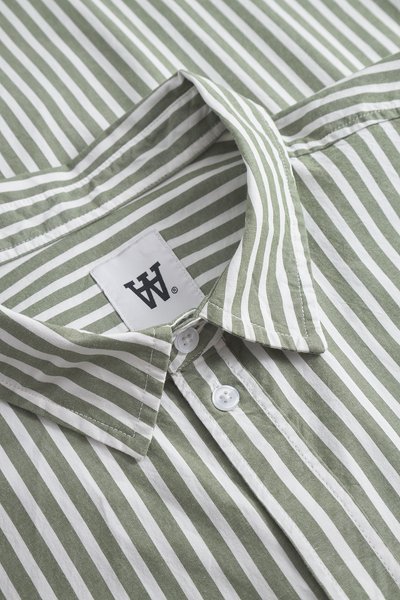 Camicia a righe verdi e bianche con colletto pieghevole, bottoni bianchi e un'etichetta in tessuto con un logo nero "WA" all'interno del colletto.