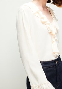 Frau in einer cremefarbenen Bluse mit Rüschensaum am Kragen und an den Ärmeln, in dunkelblauem, hochtailliertem Jeans eingesteckt, von Hals bis Hüfte gezeigt.