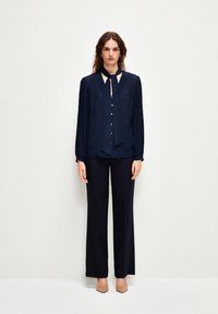 Blusa blu navy con colletto a cravatta, abbottonatura davanti, maniche lunghe plissettate e colletto a contrasto; abbinata a pantaloni a gamba larga e tacchi beige.