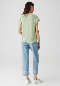 Top verde chiaro a maniche corte, vestibilità rilassata, abbinato a jeans azzurri con risvolto e sneakers bianche, visto da dietro.