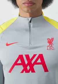 Harmaa urheilu suorituspaita korkealla kauluksella, jossa on keltaiset yksityiskohdat, punainen Nike-logo, 'L.F.C.'-embleemi ja suuri punainen 'AXA' -teksti.