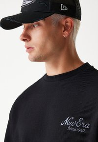 Schwarzer Sweatshirt mit strukturiertem Stoff und geripptem Material, mit gesticktem "New Era"-Logo in Weiß. Schwarze Mesh-Baseballmütze mit einem Vorderpatch und metallischen Akzenten.