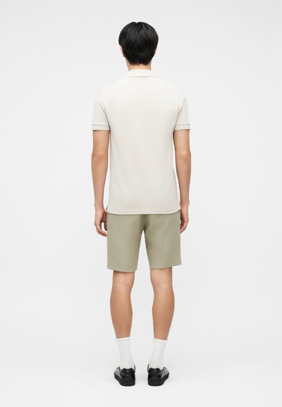 Άνδρας που στέκεται με την πλάτη στραμμένη, φοράει λευκό πόλο με κοντά μανίκια, ανοιχτό μπεζ σορτς, λευκές κάλτσες και μαύρα παπούτσια σε λευκό φόντο.