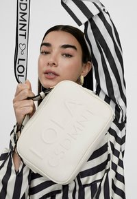 Donna con camicia a righe bianco e nere tiene una borsa bianca con rilievo del logo e una tracolla, mentre è visibile un orecchino ad anello dorato.