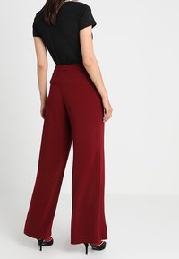 Bordeauxrode broek met wijde pijpen en een hoge tailleband, gemaakt van zachte stof, gecombineerd met een aansluitend zwart topje. Zwarte hoge hakken zichtbaar.