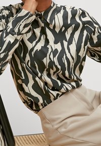 Long-sleeved blouse in black and cream with a bold abstract pattern, featuring a button collar and textured fabric. Paired with beige trousers.
