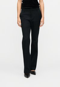 Femme portant un pantalon droit noir et des chaussures noires à bouts pointus, debout devant un fond blanc uni.