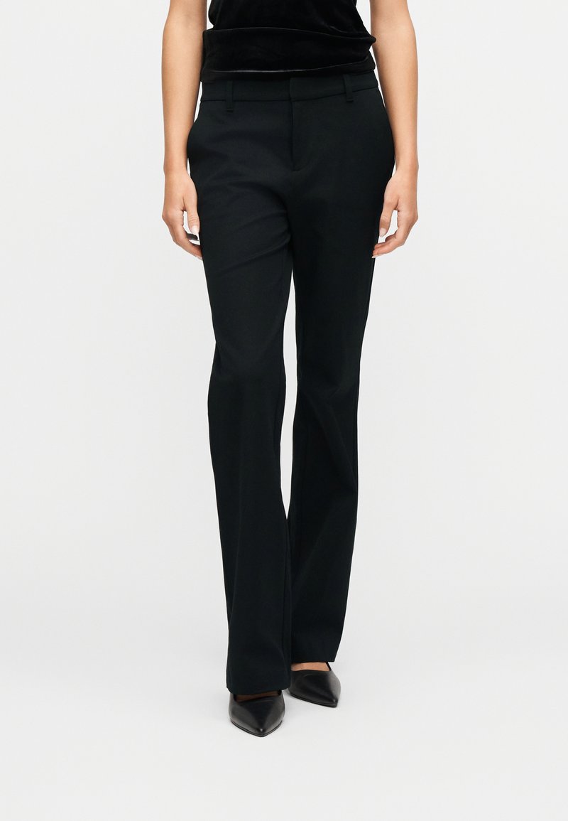 Femme portant un pantalon droit noir et des chaussures noires à bouts pointus, debout devant un fond blanc uni.