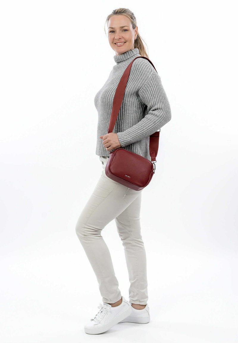 Borsa a tracolla rossa in pelle con una texture liscia, forma rettangolare e tracolla regolabile. Indossata con pantaloni attillati di colore chiaro e scarpe bianche.