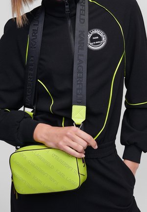 Henkilö, jolla on musta verryttelyasu, jossa on neonkeltaiset koristesaumat, ja hän pitää vaaleanvihreää Karl Lagerfeldin olkalaukkua, jossa on leveä musta merkkihihna.