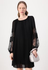 Abito nero a pieghe con maniche in pizzo trasparente e scollatura rotonda. La modella tiene una borsa in satin argento. Design semplice ed elegante con contrasto di texture.