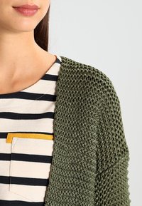 Cárdigan de punto verde oliva con patrón texturizado, colgado sobre una camiseta a rayas con un bolsillo acentuado en amarillo.