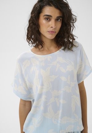 Femeie cu păr creț închis la culoare, purtând o bluză albastru deschis cu modele florale albe, stând pe un fundal simplu.