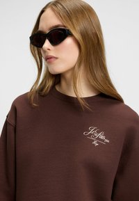 Felpa marrone con scollo a costine, caratterizzata da un testo ricamato in bianco "Je ne fais pas" e "Hugo" sul petto. Accessoriata con occhiali da sole neri a forma di occhio di gatto.