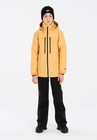 Veste imperméable jaune avec un col haut, des poches zippées et des accents noirs. Portée avec un pantalon noir et un bonnet en tricot noir.