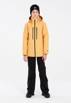 Gelbe wasserdichte Jacke mit hohem Kragen, Reißverschluss-Taschen und schwarzen Akzenten. Getragen mit schwarzen Hosen und einer schwarzen Strickmütze.