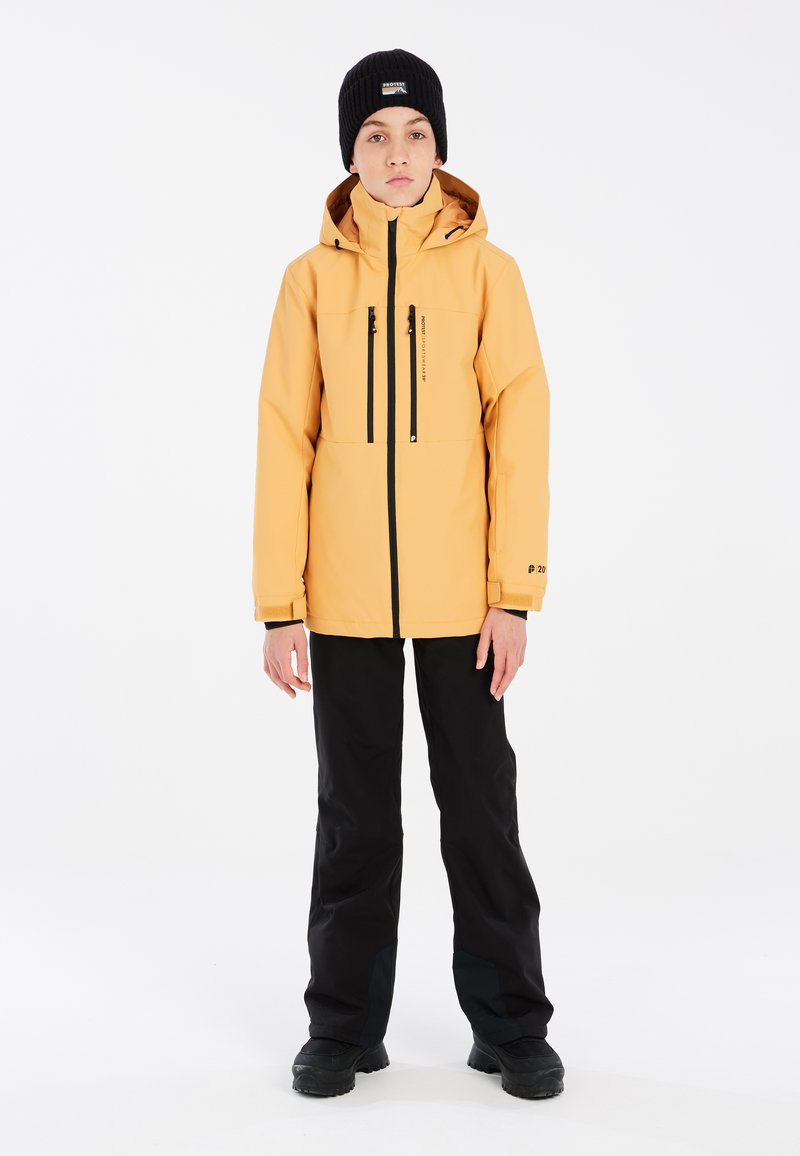 Veste imperméable jaune avec un col haut, des poches zippées et des accents noirs. Portée avec un pantalon noir et un bonnet en tricot noir.
