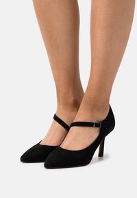 Nohy ženy obuté do černých semišových Mary Jane lodiček s kotníkovými pásky a špičatou špičkou na bílém pozadí.
