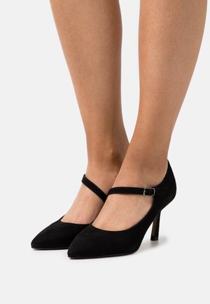 Nohy ženy obuté do černých semišových Mary Jane lodiček s kotníkovými pásky a špičatou špičkou na bílém pozadí.