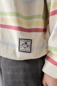Prúžkovaná mikina v krémovej farbe so svetlo modrými, zelenými, žltými a červenými pruhmi. Obsahuje štvorcový nášivku s textom "KAOTIKO SOCIETY" a grafikou.