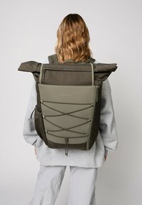 Zaino a mazzetta verde oliva con tessuto testurizzato e dettaglio con corda elastica. Presenta spallacci regolabili e scomparti con zip.