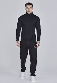 Black turtleneck shirt, fitted design; paired with black cargo pants featuring pockets and gold embroidered detail; white sneakers.