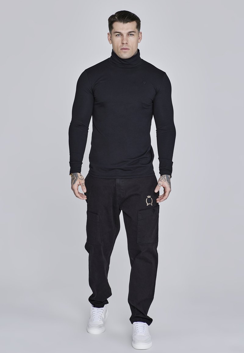 Black turtleneck shirt, fitted design; paired with black cargo pants featuring pockets and gold embroidered detail; white sneakers.