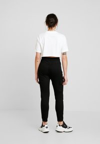 Pantalones cargo negros con bolsillos laterales, combinados con una camiseta blanca corta. Modelo usando zapatillas negras y blancas, de espaldas.
