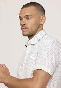 Chemise blanche à manches courtes avec un motif en losange bleu marine ; col boutonné ; tissu léger ; couture visible ; modèle vu de profil.