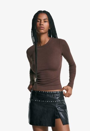 Bruine lange mouwen aansluitende top, gecombineerd met een zwarte leren minirok met zilveren studs en een zijsplit, geaccentueerd met een riem.