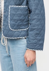 Άτομο που φοράει μπλε καπιτονέ μπουφάν με μεγάλη τσέπη και περιγράμματα, συνδυασμένο με ανοιχτόχρωμο τζιν παντελόνι και λευκό πουκάμισο με φαρδιά κορδόνια.