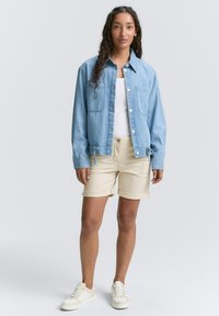 Veste en denim bleu clair avec deux poches poitrine, portée sur un haut blanc ; associée à un short beige retroussé et des baskets blanches.