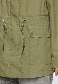 Veste olive en vert avec un cordon de serrage à la taille, poche à bouton sur le devant et texture de tissu lisse avec des détails de couture visibles.