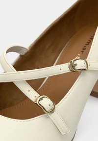 Λευκό δερμάτινο παπούτσι με σχέδιο σταυρωτής ζώνης και δύο χρυσές αγκράφες. Απαλή υφή με ελαφρώς στρογγυλή μύτη και χαμηλό τακούνι.