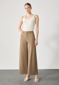 Beige nátělník spárovaný s hnědými kalhotami s vysokým pasem a širokými nohavicemi. Modelka stojí proti jednobarevnému pozadí a má na sobě béžové sandály na podpatku s pásky.