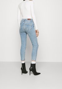 Jeans skinny azul claro con cintura alta, que presentan un detalle desgastado en la pierna derecha y sutiles acentos de cremallera en el tobillo.