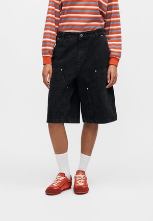 Person iført sorte knælange shorts med store sidelommer, hvide sokker, røde sneakers og en orange, blå og hvide stribet langærmet trøje.