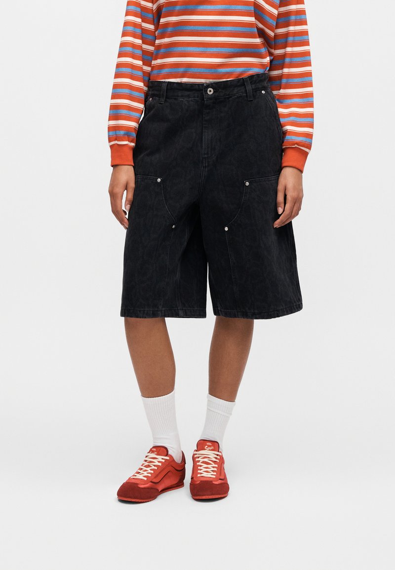 Person trägt schwarze knielange Shorts mit großen Seitentaschen, weiße Socken, rote Sneakers und ein orange-blau-weiß gestreiftes Langarmshirt.