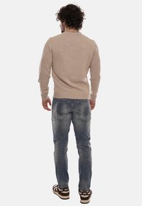 Uomo con capelli ricci che indossa un maglione beige, jeans blu strappati e sneakers Nike marroni, in piedi rivolto verso il retro su uno sfondo bianco.