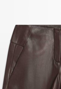 Pantaloni in pelle marrone con una texture liscia, dotati di tasche laterali e un taglio su misura, che evidenziano un design minimalista e materiali di qualità.