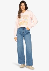 Helles rosa Sweatshirt mit Grafikdruck, kombiniert mit hochgeschnittenen, blauen weit geschnittenen Jeans und beigen Schuhen. Der Stoff wirkt weich; lässiges Design.